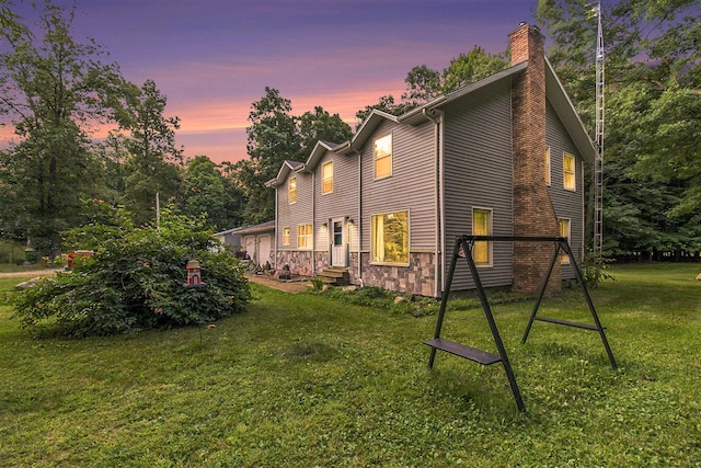 back of property at dusk featuring a garage, stone siding, a chimney, and a lawn