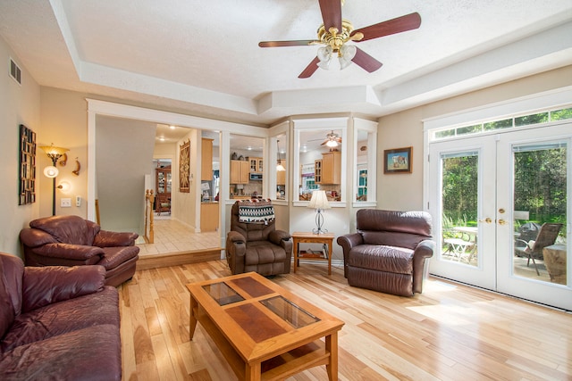 living room featuring ceiling fan, a textured ceiling, a tray ceiling, light hardwood / wood-style flooring, and french doors