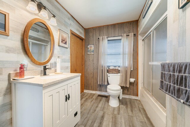 full bathroom with wood-type flooring, shower / bath combination with glass door, wooden walls, vanity, and toilet