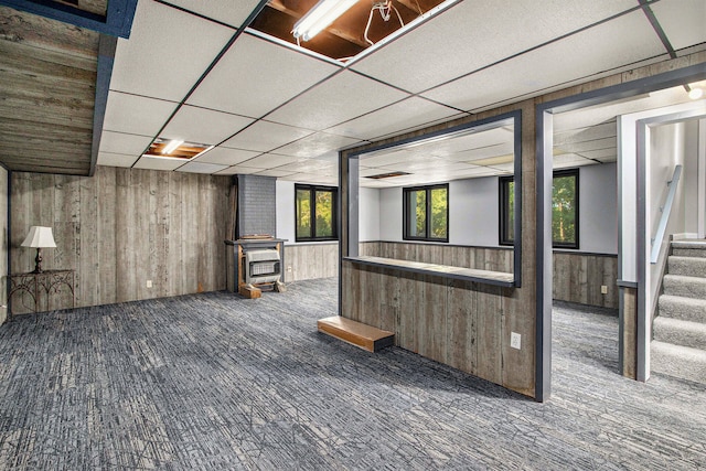 basement with carpet floors, a drop ceiling, a wood stove, and wooden walls