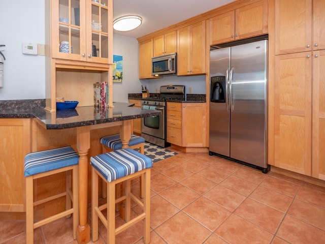 kitchen with dark stone countertops, a kitchen bar, light brown cabinetry, appliances with stainless steel finishes, and light tile patterned flooring