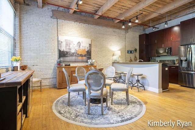 dining room featuring rail lighting, beamed ceiling, light hardwood / wood-style flooring, and brick wall