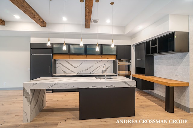 kitchen with beam ceiling, a kitchen island with sink, decorative light fixtures, and light wood-type flooring