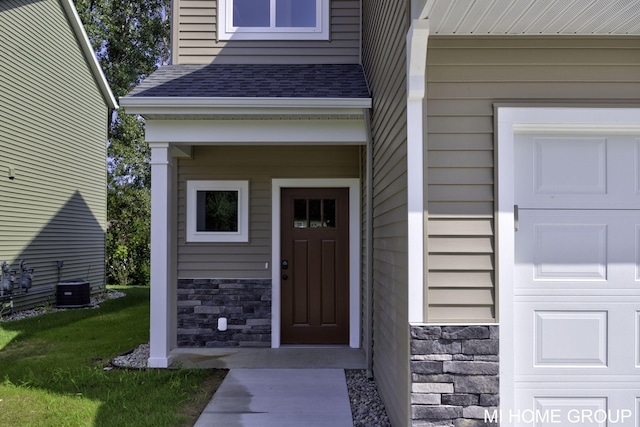 property entrance with central air condition unit, stone siding, a yard, roof with shingles, and a garage