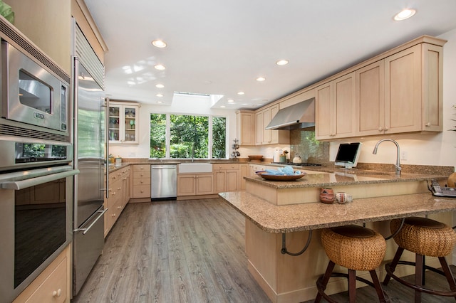 kitchen with built in appliances, light hardwood / wood-style flooring, a skylight, kitchen peninsula, and wall chimney range hood