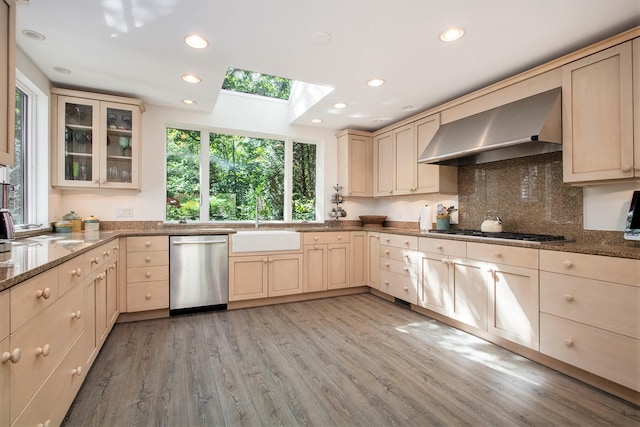 kitchen with stone countertops, a skylight, wall chimney exhaust hood, appliances with stainless steel finishes, and light hardwood / wood-style floors