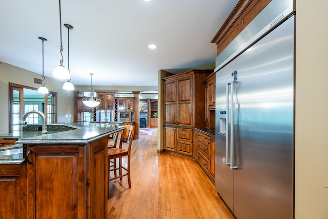 kitchen featuring a kitchen island with sink, light wood-type flooring, built in refrigerator, a kitchen breakfast bar, and hanging light fixtures