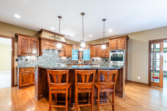 kitchen featuring a kitchen island with sink, light wood-type flooring, stainless steel oven, and hanging light fixtures