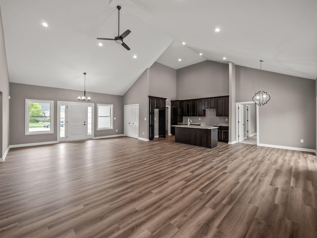 unfurnished living room featuring high vaulted ceiling, ceiling fan with notable chandelier, a sink, wood finished floors, and baseboards