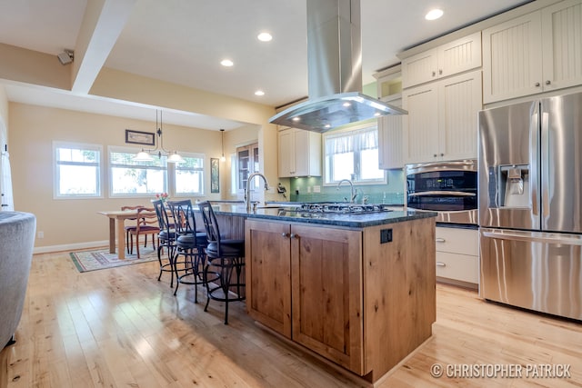 kitchen with dark stone countertops, light hardwood / wood-style flooring, island exhaust hood, an island with sink, and appliances with stainless steel finishes