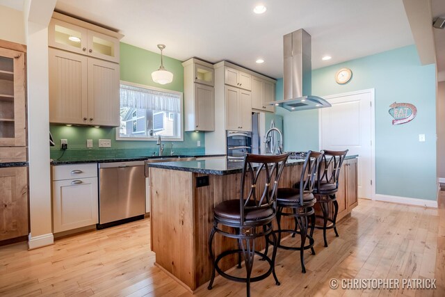 kitchen featuring decorative light fixtures, a kitchen island with sink, island exhaust hood, stainless steel appliances, and light wood-type flooring