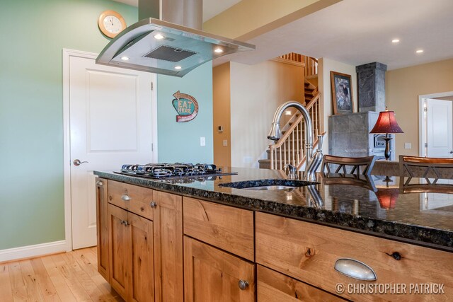 kitchen with dark stone countertops, light hardwood / wood-style flooring, sink, stainless steel gas cooktop, and island range hood