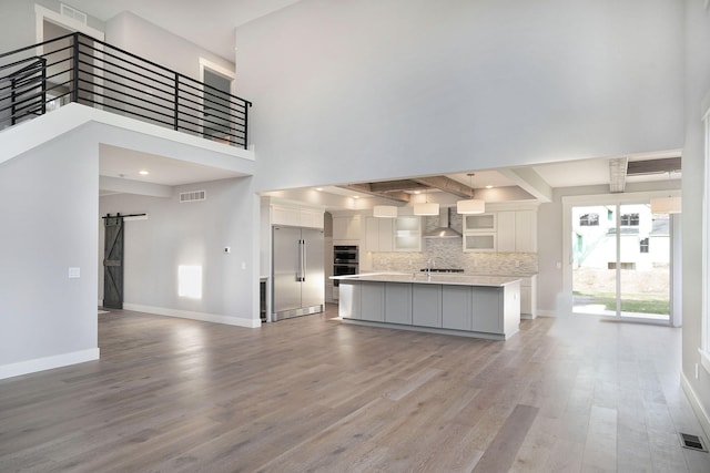 kitchen featuring a kitchen island with sink, tasteful backsplash, white cabinets, a barn door, and wall chimney exhaust hood