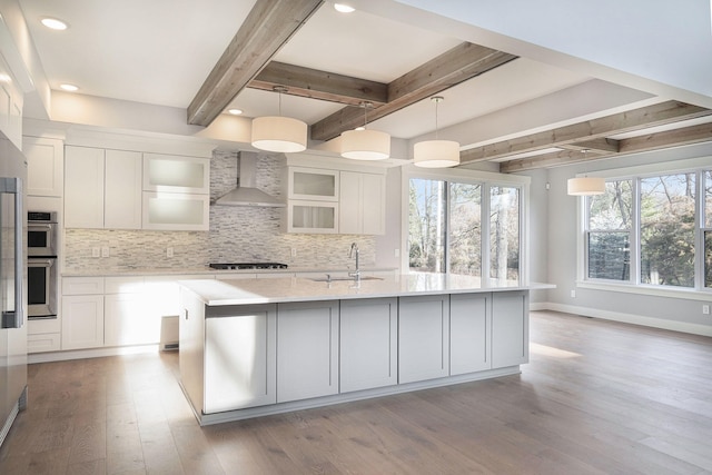 kitchen with decorative light fixtures, sink, white cabinets, wall chimney range hood, and a center island with sink