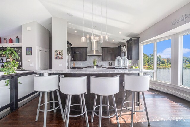 kitchen featuring a large island with sink, vaulted ceiling, backsplash, dark brown cabinets, and wall chimney exhaust hood
