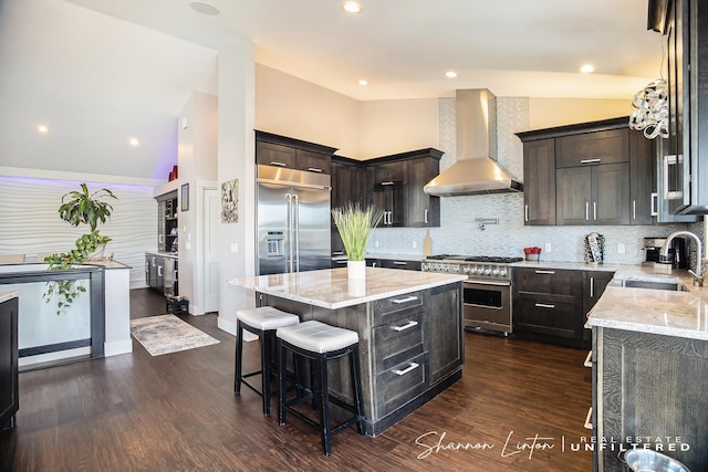 kitchen with lofted ceiling, a sink, light stone countertops, premium appliances, and wall chimney exhaust hood