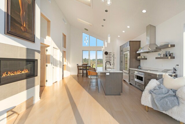 kitchen with island range hood, a tiled fireplace, stainless steel oven, light hardwood / wood-style flooring, and a kitchen island with sink