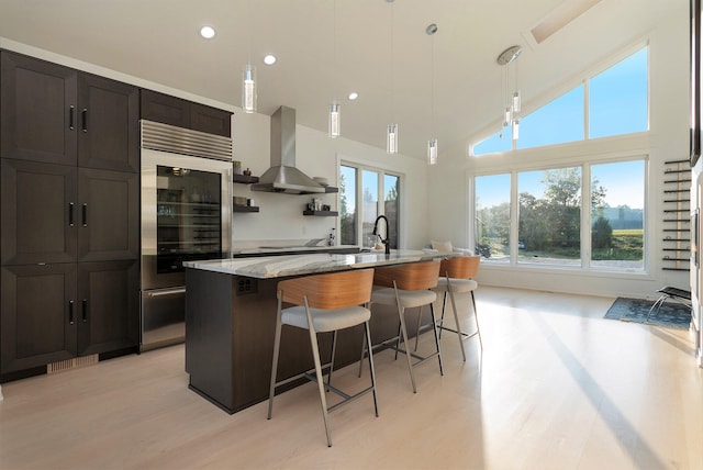 kitchen with a towering ceiling, wall chimney exhaust hood, light stone countertops, pendant lighting, and a center island with sink