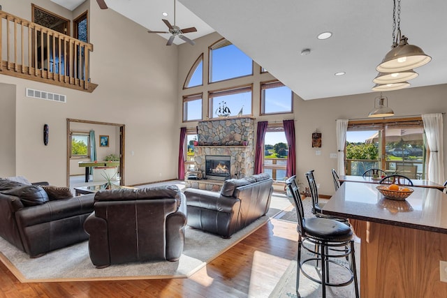 living room featuring a towering ceiling, light wood-type flooring, a stone fireplace, a ceiling fan, and visible vents