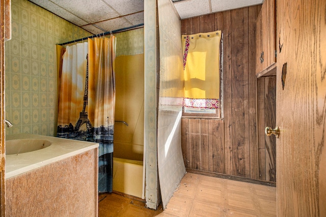 bathroom featuring shower / bath combination with curtain, wood walls, a drop ceiling, and sink