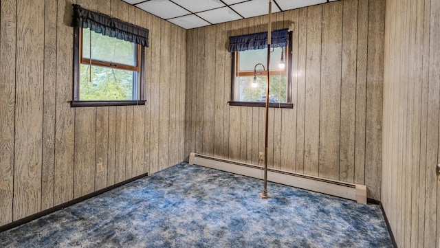 unfurnished room with dark carpet, wooden walls, a drop ceiling, and a baseboard heating unit