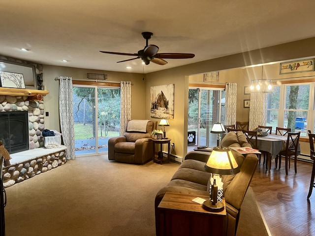 living room featuring ceiling fan with notable chandelier, a healthy amount of sunlight, a stone fireplace, and a baseboard heating unit