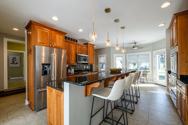 kitchen with ceiling fan, an island with sink, hanging light fixtures, backsplash, and stainless steel appliances