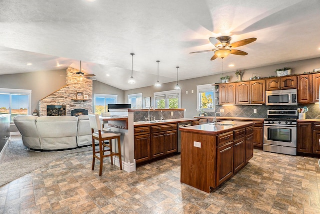 kitchen featuring appliances with stainless steel finishes, hanging light fixtures, vaulted ceiling, a stone fireplace, and a center island with sink