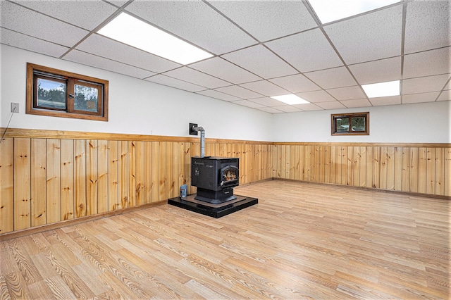 basement featuring light hardwood / wood-style floors, a paneled ceiling, wooden walls, and a wood stove