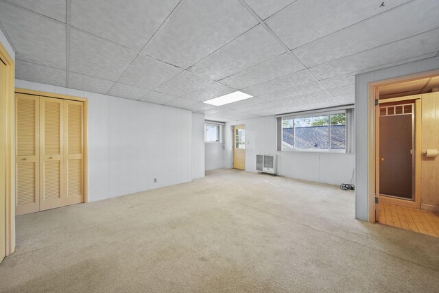 basement with a drop ceiling, heating unit, light colored carpet, and wooden walls