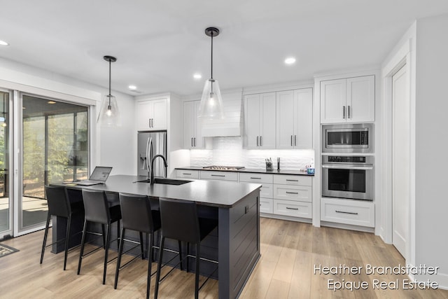 kitchen with white cabinets, an island with sink, decorative light fixtures, stainless steel appliances, and light hardwood / wood-style floors