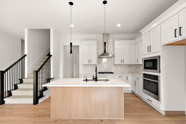 kitchen with wall chimney range hood, oven, an island with sink, sink, and white cabinetry