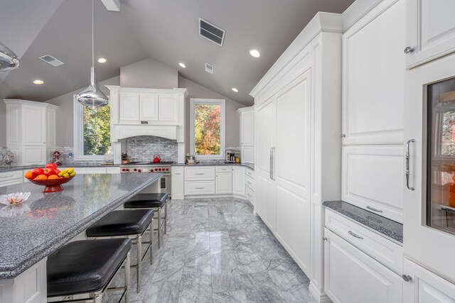 kitchen with dark stone countertops, high end stainless steel range oven, white cabinetry, vaulted ceiling, and a breakfast bar area