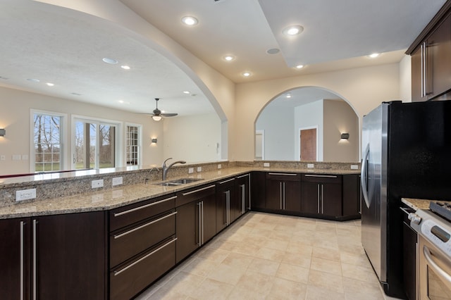 kitchen with dark brown cabinetry, ceiling fan, sink, stainless steel appliances, and light stone countertops