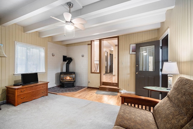 living room with ceiling fan, beamed ceiling, a wood stove, light hardwood / wood-style flooring, and wooden walls