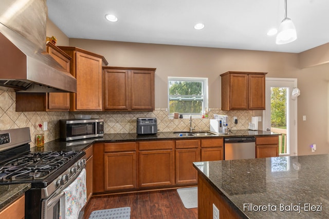 kitchen with wall chimney range hood, dark hardwood / wood-style flooring, appliances with stainless steel finishes, dark stone counters, and decorative light fixtures