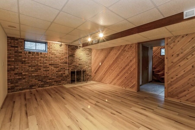 basement with hardwood / wood-style floors, a fireplace, a drop ceiling, wooden walls, and brick wall