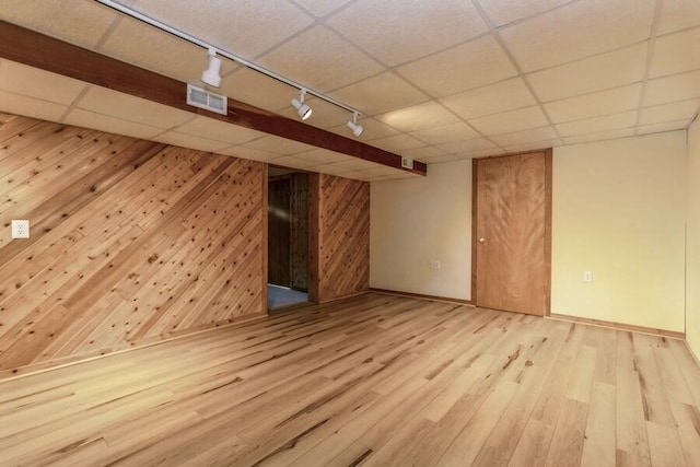 basement featuring rail lighting, light hardwood / wood-style floors, a paneled ceiling, and wooden walls