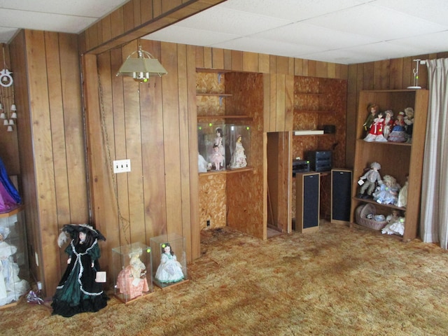 interior space with carpet floors, a drop ceiling, hanging light fixtures, and wooden walls