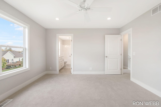 unfurnished bedroom featuring ensuite bathroom, multiple windows, light colored carpet, and ceiling fan