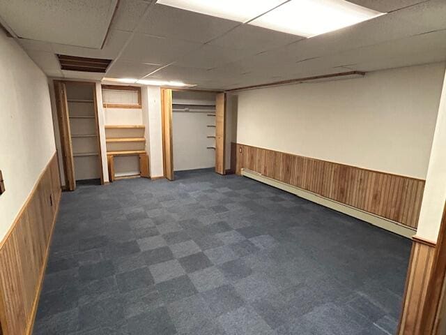 basement with a paneled ceiling, a baseboard radiator, wooden walls, and dark carpet