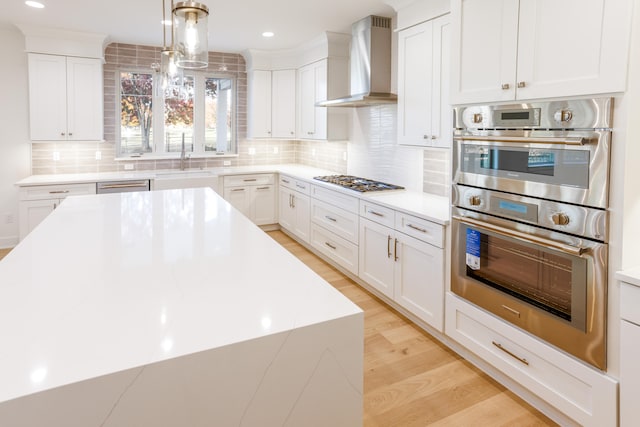 kitchen with stainless steel appliances, hanging light fixtures, white cabinets, wall chimney range hood, and light wood-type flooring