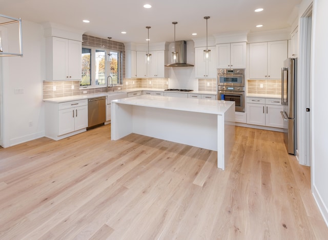 kitchen with stainless steel appliances, a center island, wall chimney range hood, white cabinetry, and decorative light fixtures