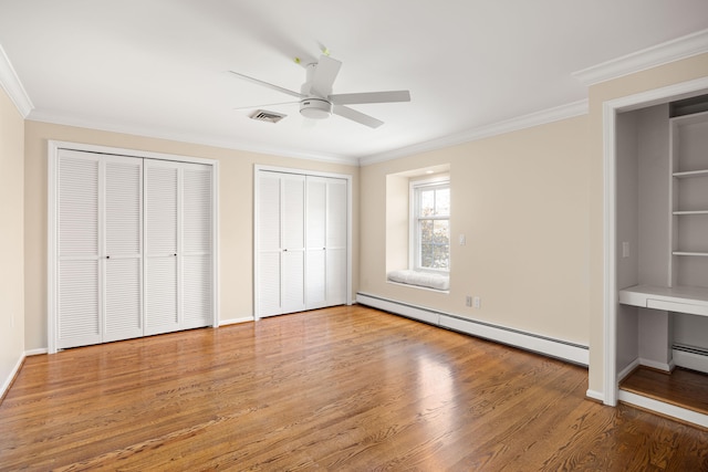 unfurnished bedroom featuring a baseboard heating unit, hardwood / wood-style flooring, ornamental molding, ceiling fan, and multiple closets