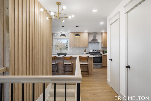 kitchen featuring wall chimney range hood, decorative light fixtures, light brown cabinets, stainless steel range oven, and light hardwood / wood-style flooring