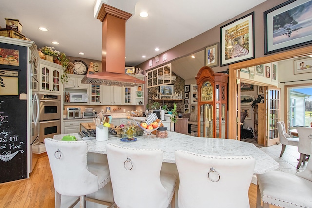 kitchen featuring light stone countertops, a kitchen bar, island range hood, stainless steel double oven, and light hardwood / wood-style flooring