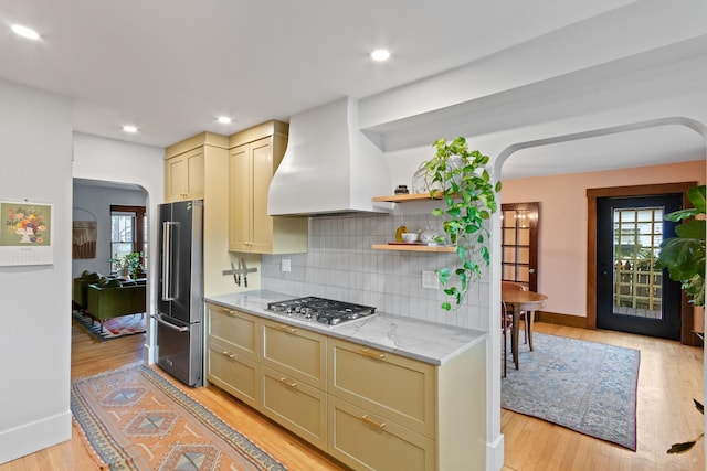 kitchen featuring tasteful backsplash, light stone counters, custom exhaust hood, stainless steel appliances, and cream cabinetry