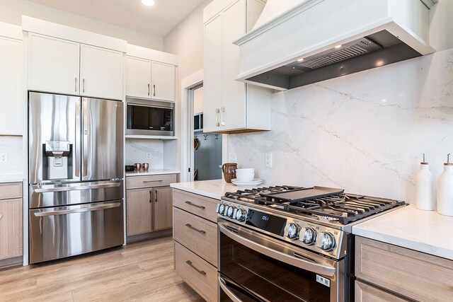 kitchen with light hardwood / wood-style floors, appliances with stainless steel finishes, white cabinets, premium range hood, and decorative backsplash