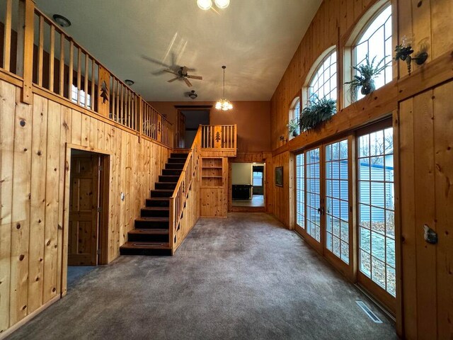 unfurnished living room featuring a high ceiling, ceiling fan with notable chandelier, dark colored carpet, and wooden walls