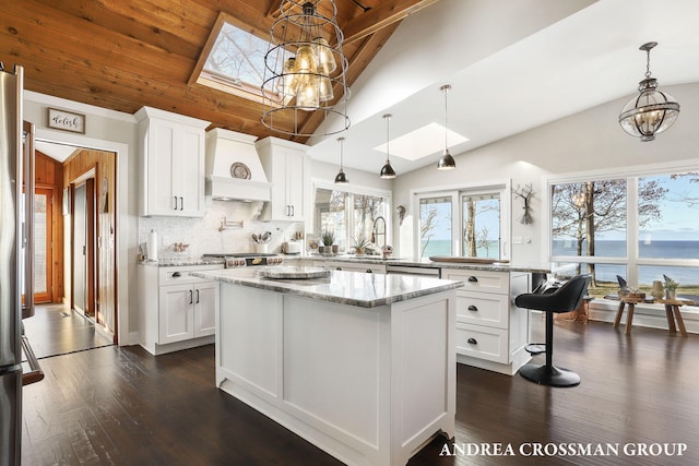 kitchen with light stone counters, custom exhaust hood, lofted ceiling with skylight, a water view, and white cabinetry